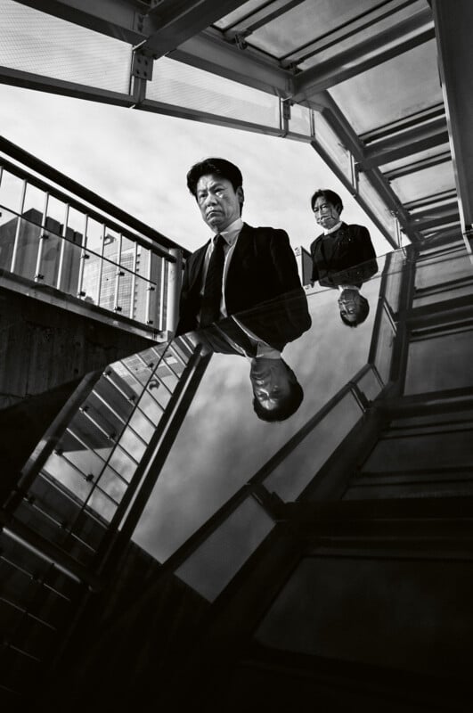 Two people in suits stand on a staircase under a glass roof, with their reflections visible on a shiny surface below. The image is in black and white, creating a dramatic, moody atmosphere.