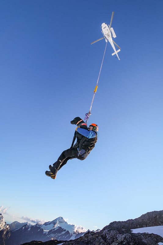 A person in outdoor gear is being airlifted by a helicopter, suspended high above snowy mountains under a clear blue sky.