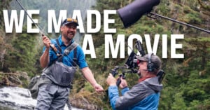 Two men stand in a forested area by a river; one holds a fishing rod and laughs while the other films him with a camera. Large text in the background reads, "WE MADE A MOVIE.