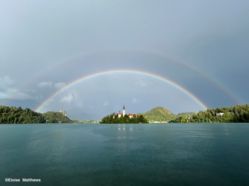 Un doble arcoiris se curva sobre un lago tranquilo con una isla en la isla con una iglesia en el centro rodeada de colinas verdes bajo un cielo nublado.