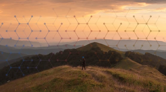 A person stands on a grassy hilltop at sunset with rolling hills and a colorful sky in the background; a blue and orange geometric pattern overlays the image.
