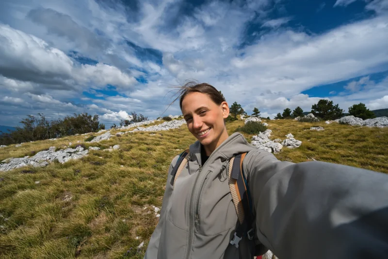 Una mujer con una chaqueta gris tomando fotos mientras camina en una colina nublada parte de una caminata. Árboles y arbustos bajos en el backstage.