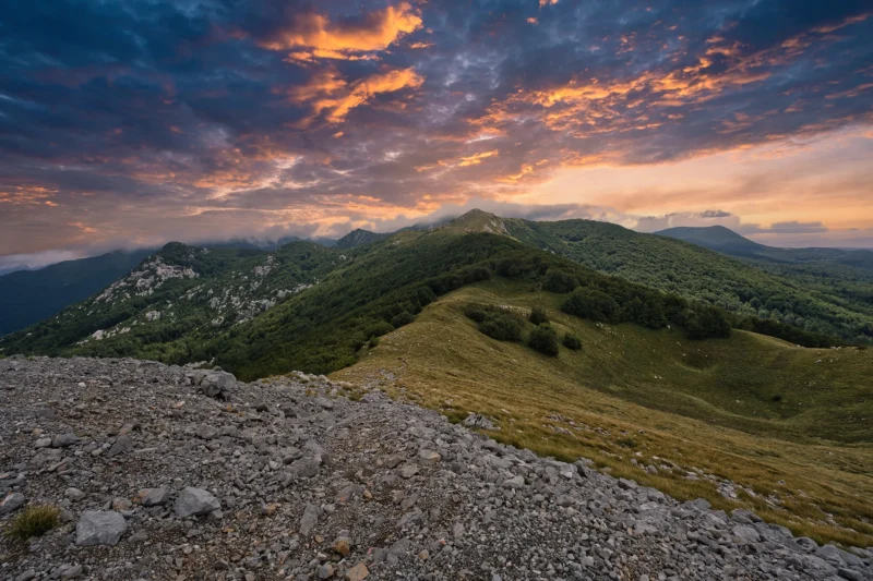 El primer plano rocoso da a las colinas verdes y montañas boscosas, bajo el dramático cielo de puesta de sol con vibrantes nubes de naranja y púrpura.
