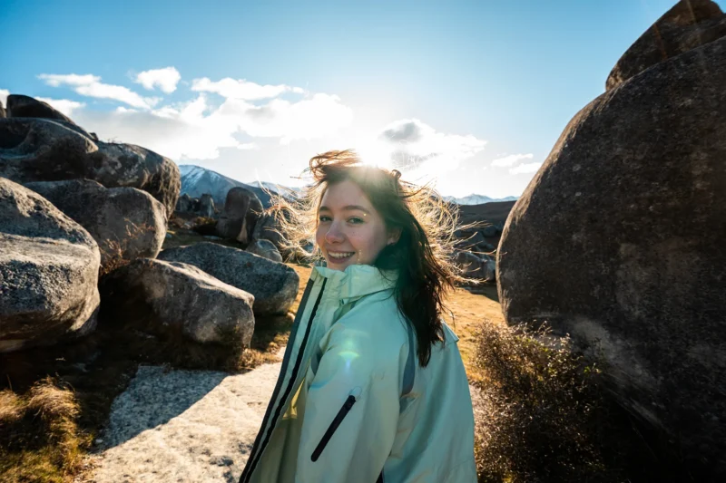 Una mujer joven con una chaqueta ligera sonríe a la cámara mientras está de pie en una enorme roca al aire libre. El sol brilla detrás de ella, resaltando su cabello con cielo azul y nubes dispersas.