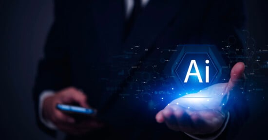 A person in a suit holds a glowing hexagonal icon with the letters "AI" above their open palm, representing artificial intelligence, while using a smartphone in the other hand. The background is dark and tech-themed.