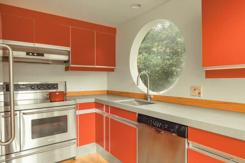 Cocina moderna con gabinetes rojos, electrodomésticos de acero inoxidable, encimeras grises y ventanas redondas sobre el fregadero con vistas a la vegetación. Una pequeña olla roja se encuentra en la estufa.