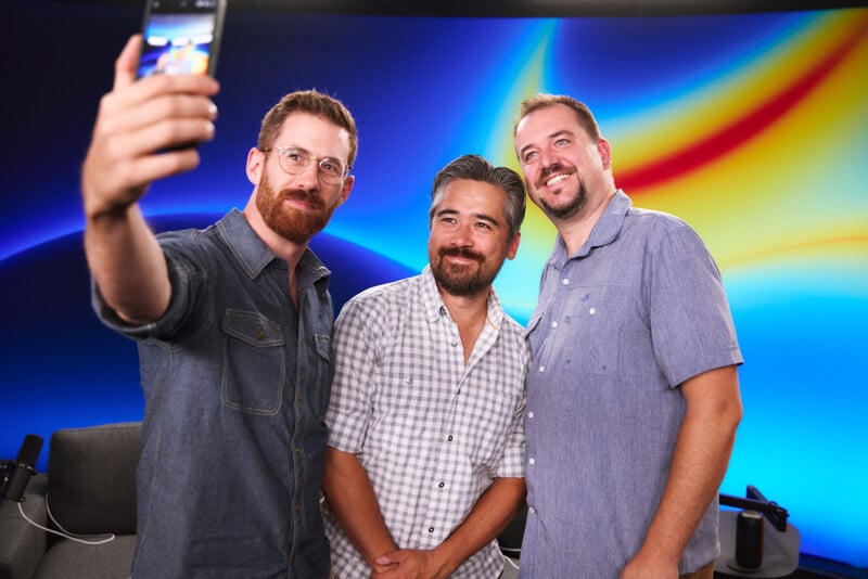 Three men in casual shirts smile as one takes a selfie with his smartphone. They stand in front of a colorful, abstract background with a blue gradient and yellow and red streaks. Microphones are visible in the foreground.
