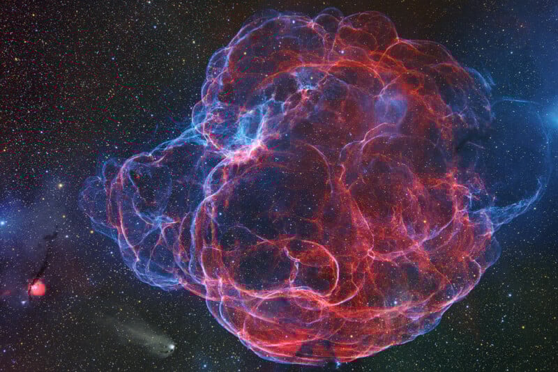 A colorful, intricate nebula with glowing red and blue filaments expands against a star-filled background in deep space. The nebula appears like a tangled, luminous cloud with varying intensity and detail.