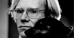 A black-and-white photo of a person with light hair and glasses holding a dark-colored dog close to their face, looking directly at the camera.