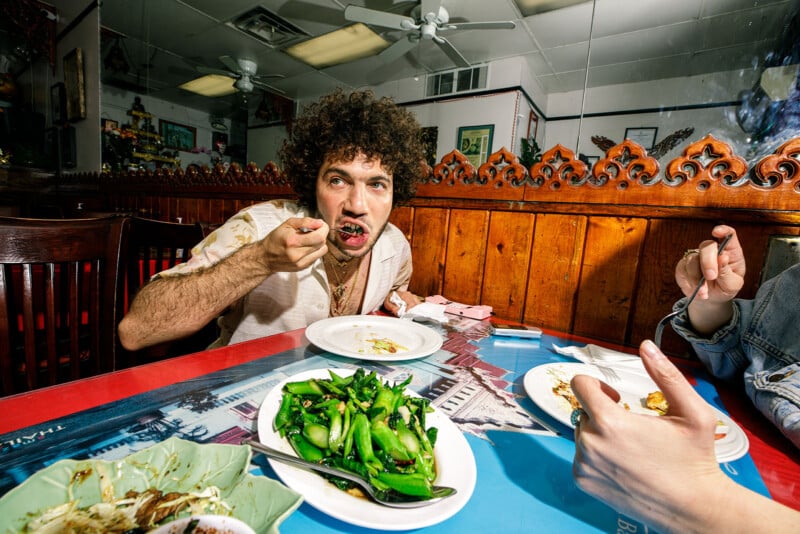 Un hombre con cabello rizado se come en la mesa de un restaurante y se pone su tenedor en la boca. Vegetales verdes y platos de comida frente a él. Otra persona es parcialmente visible y puede obtener comida. Establezca brillante y colorido.