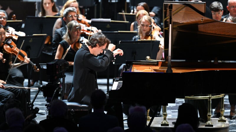 A pianist passionately plays a grand piano on stage, surrounded by an orchestra of string musicians. The scene captures a live classical music performance with attentive musicians and an engaged audience.