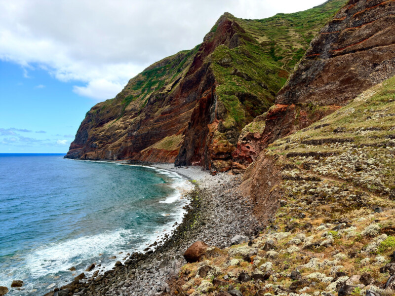 Rocky coastline with steep, green and reddish-brown cliffs beside the blue ocean under a partly cloudy sky, with waves gently hitting the stony shore.