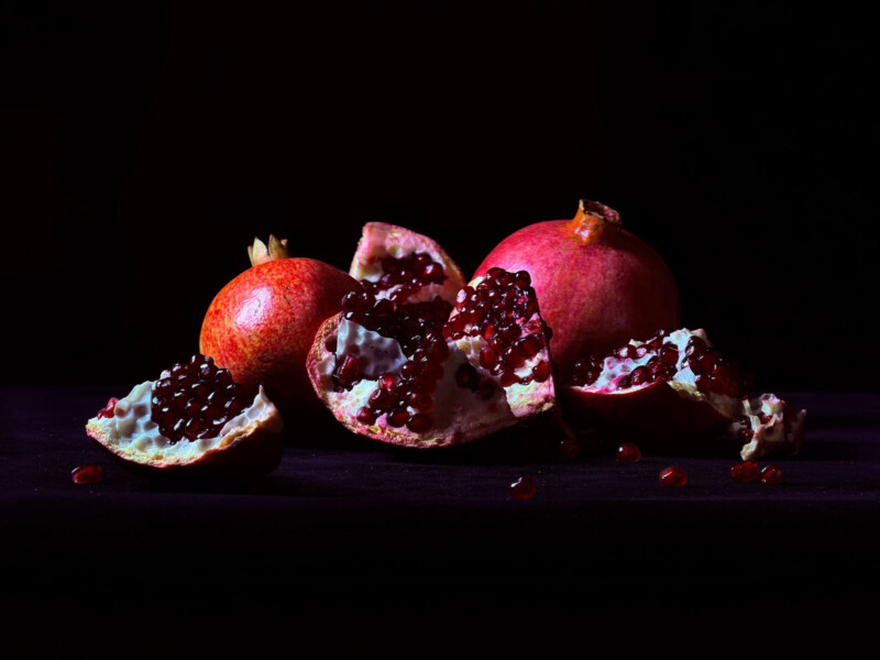 Two whole pomegranates and several pomegranate halves with glistening red seeds are displayed on a dark surface against a black background.