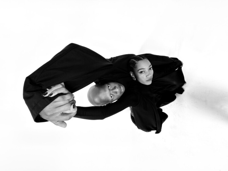 A bald man and a woman in black outfits hold hands and gaze upward, shot from above against a white background, creating a dramatic, artistic composition.