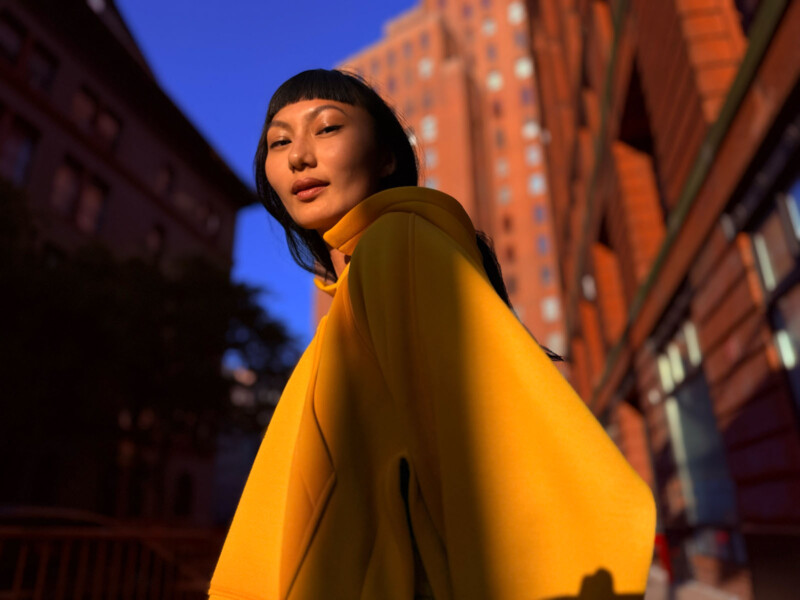 A person wearing a bright yellow coat stands outdoors in a city, with tall brick buildings in the background. Warm sunlight illuminates their face and clothing, creating strong shadows and a dramatic effect.
