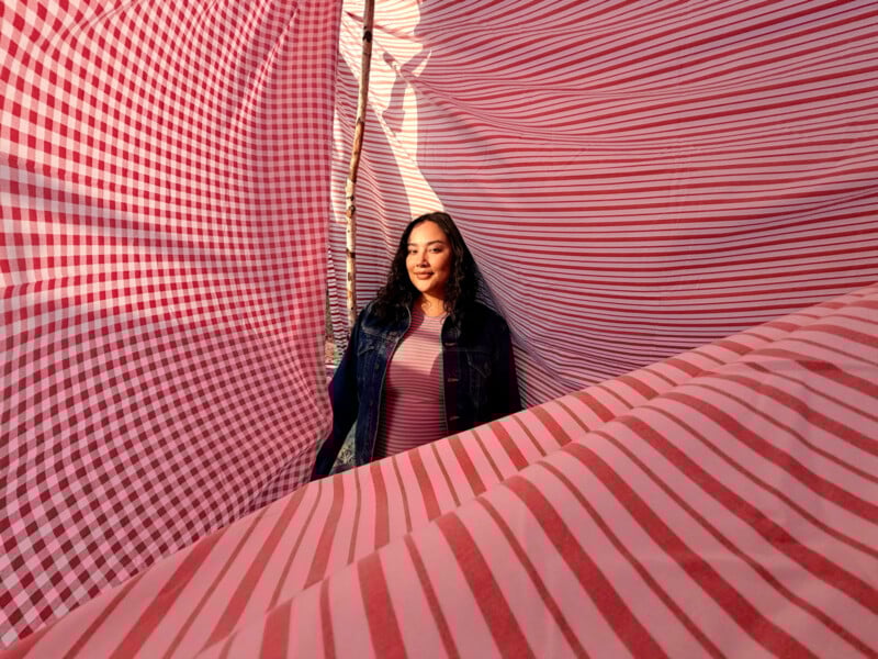 A woman in a denim jacket and pink shirt stands surrounded by large red and white gingham-patterned fabric sheets, smiling softly as sunlight filters through the material.