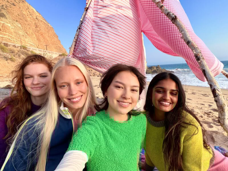 Four young women smile and pose for a selfie on a sandy beach, with a striped pink fabric tent and ocean waves in the background. They are wearing bright, colorful sweaters.