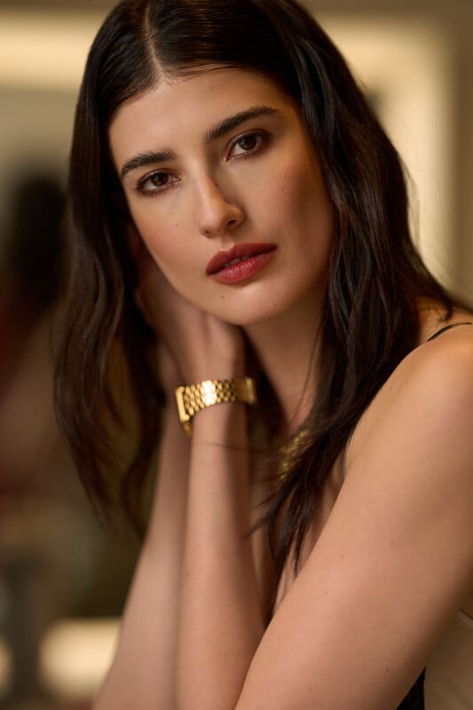 A woman with long dark hair and red lipstick gazes softly at the camera, resting her hands near her neck. She wears a gold watch and a sleeveless top, with a softly blurred background.