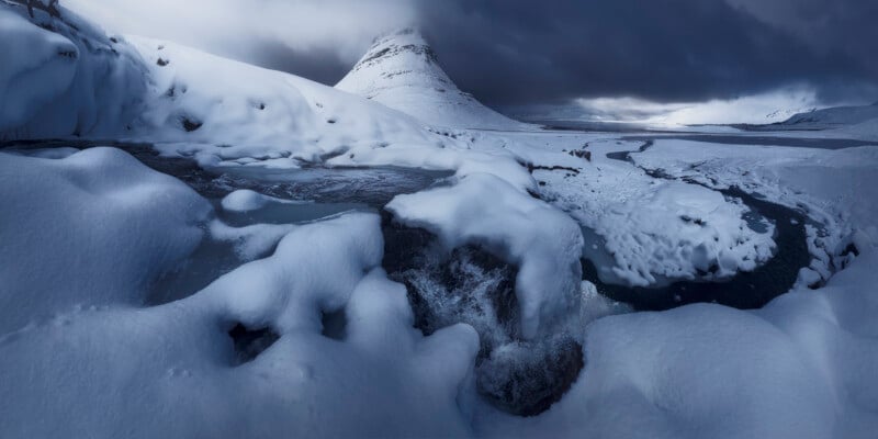 A snow-covered landscape with a mountain in the background, dark clouds overhead, and a partially frozen river winding through the snowy terrain in the foreground.