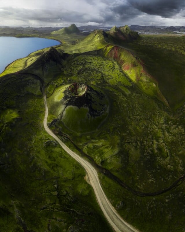 A winding road cuts through lush, green volcanic hills under a cloudy sky, with a crater mound in the foreground and a blue lake visible to the left. Dramatic sunlight highlights the vibrant landscape.