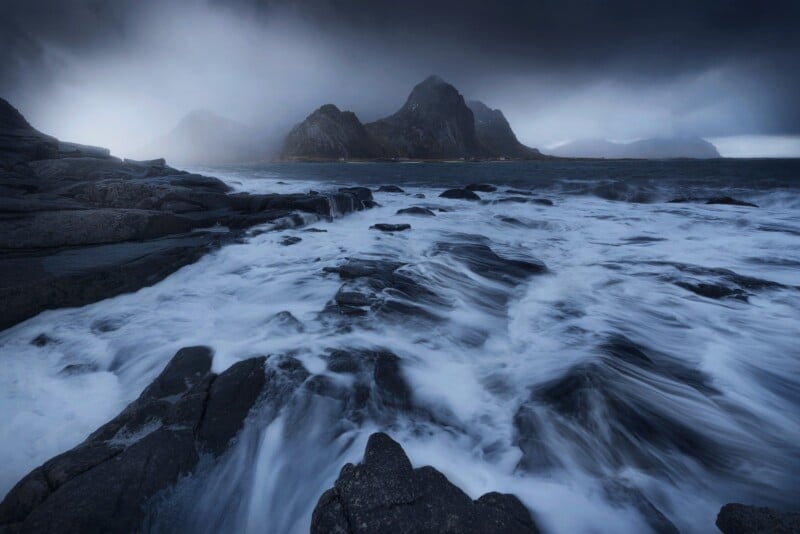 Waves crash over dark rocky shoreline under a dramatic cloudy sky, with rugged mountains in the background partly shrouded in mist.