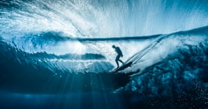 A surfer rides inside a translucent blue wave, surrounded by shimmering light rays that filter through the water, creating a dramatic and dynamic underwater perspective.