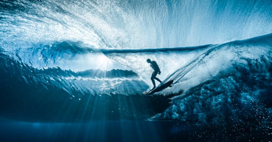 A surfer rides inside a translucent blue wave, surrounded by shimmering light rays that filter through the water, creating a dramatic and dynamic underwater perspective.