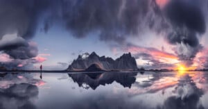 A dramatic landscape with mountains reflected in still water at sunset, clouds fill the sky, and a lone person in red stands to the left, emphasizing the vastness and beauty of the scene.
