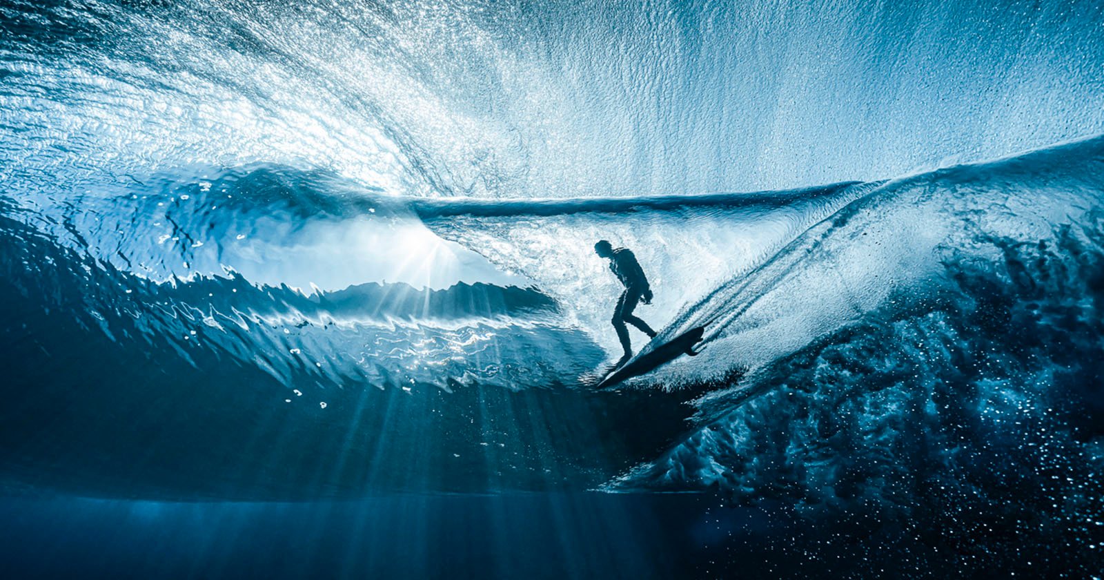 A surfer rides inside a translucent blue wave, surrounded by shimmering light rays that filter through the water, creating a dramatic and dynamic underwater perspective.