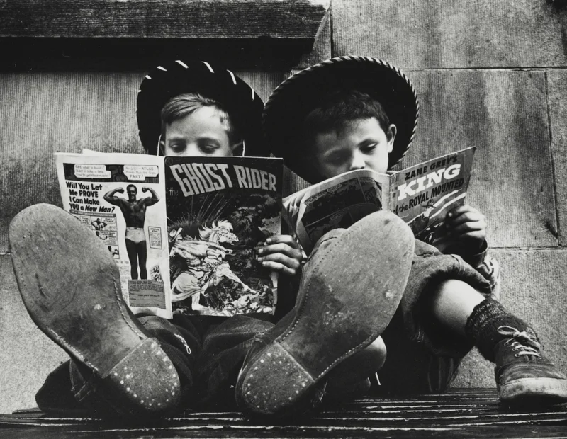 Two boys wearing hats sit side by side on a bench with their feet up, reading comic books. The comic covers are visible, and their large shoes are prominent in the foreground. The scene appears vintage and playful.