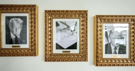 Three ornate gold-framed photos hang on a white wall; two black-and-white portraits of the same man in a suit flank a signed photo of a pen resting on paper in the center.