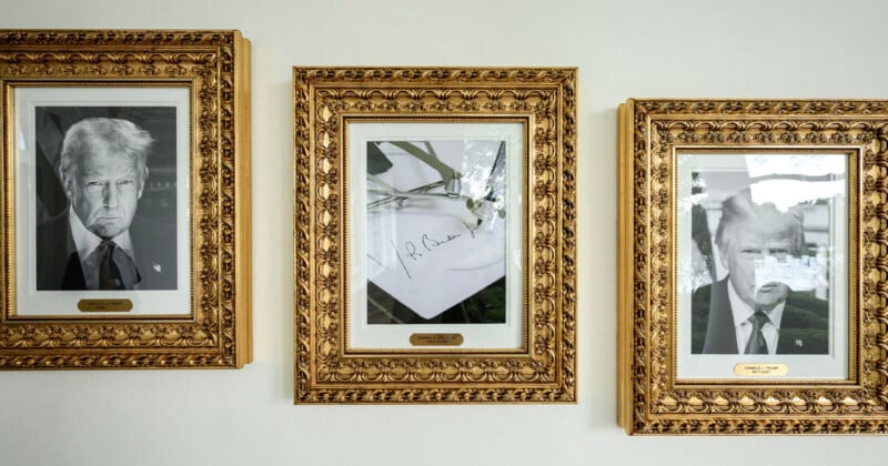 Three ornate gold-framed photos hang on a white wall; two black-and-white portraits of the same man in a suit flank a signed photo of a pen resting on paper in the center.