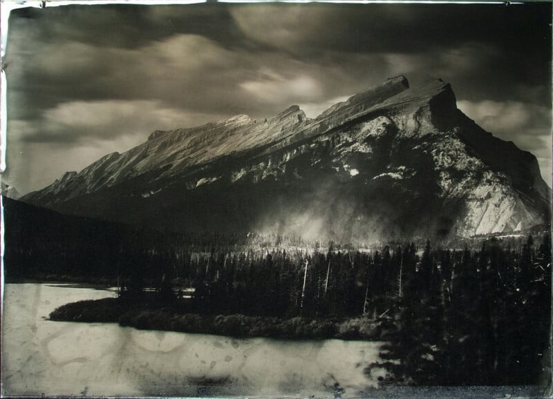 Black-and-white photo of a dramatic, jagged mountain rising above a dense pine forest and a calm river or lake, with clouds streaking across the sky, creating a moody and timeless atmosphere.