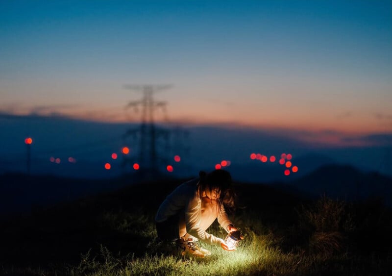 A person crouches on grassy ground at dusk, illuminated by a lantern they hold. Distant power lines and red lights are visible against a darkening blue and orange sky.