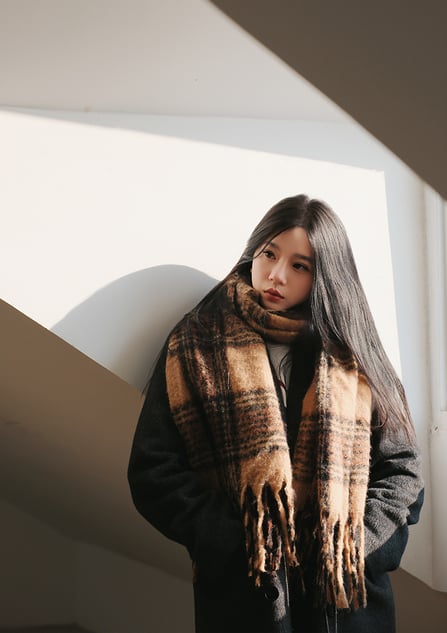 A young woman with long dark hair stands indoors, wearing a large brown plaid scarf and a dark coat. Sunlight streams through a window, casting soft shadows on the wall behind her. She gazes thoughtfully to the side.