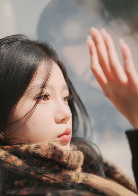 A young woman with long dark hair and a leopard-print scarf gazes thoughtfully out a window, her hand raised as if touching the glass. Her face and hand are softly lit, and her reflection is visible in the glass.