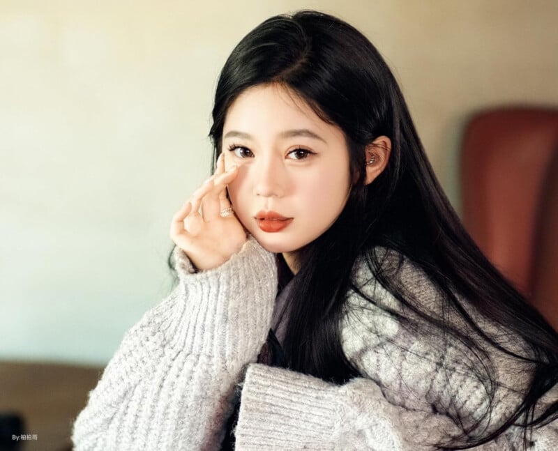 A young woman with long black hair wears a chunky gray sweater and rests her hand gently on her cheek, looking softly at the camera with a neutral background behind her.