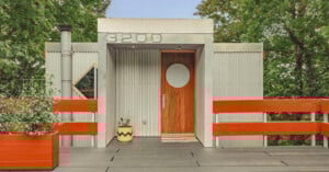 Modern house entrance with gray corrugated metal exterior, a wooden door featuring a circular window, red railings, large house number 3200 above, and potted plants by the door, surrounded by green trees.