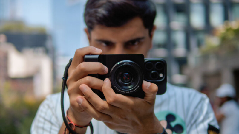 A person is holding a camera up to their face, preparing to take a photo outdoors. The background is blurred, showing buildings and greenery. The focus is on the camera and the person's hands.