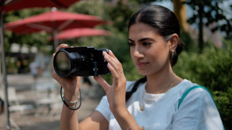 A woman with dark hair in a ponytail holds a camera, aiming it forward outdoors. She is wearing a white shirt and standing near red umbrellas and greenery on a sunny day.