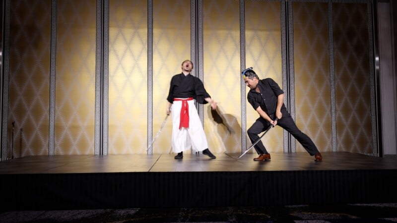 Two people performing on stage: one in a black top and white pants with a red sash, holding a sword and shouting; the other in dark clothing, crouched with a sword, facing the first person. Gold patterned background.