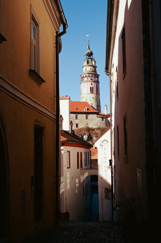 A narrow cobblestone alley between tall buildings leads to a sunlit town with a tall, ornate clock tower in the background under a clear blue sky.