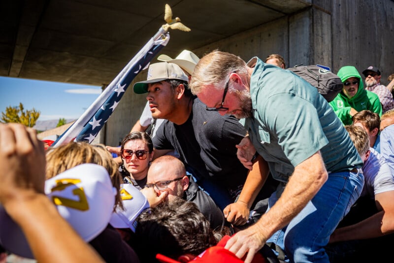A group of people are crowded together under a concrete bridge, some looking distressed. One man holds a large American flag with a gold eagle on top. The scene appears chaotic and intense.