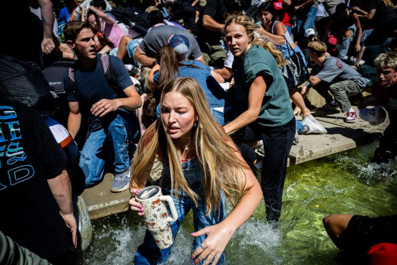 A crowd of people, some alarmed, scramble through a shallow fountain. A blond woman in overalls, holding a cup, moves through the water while others climb out, all appearing urgent and distressed.