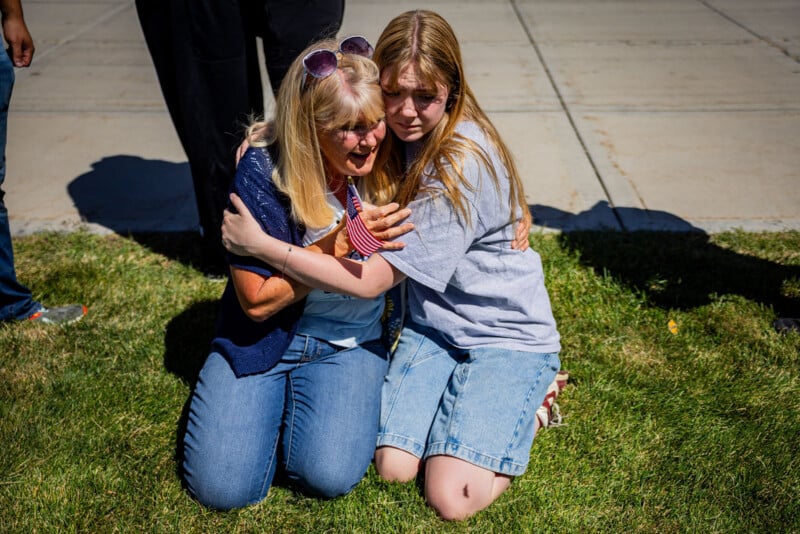 Two people kneel on the grass, hugging each other tightly. Both appear emotional, with pained expressions, as sunlight casts strong shadows. Other people’s legs are visible nearby.