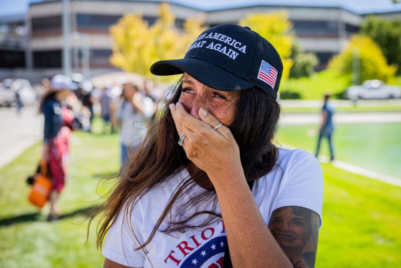 A woman wearing a "Make America Great Again" hat and a patriotic shirt stands outdoors, covering her mouth with her hand. She appears emotional. People and a building are visible in the blurred background.