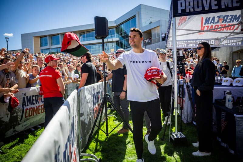 A man in a white “FREEDOM” shirt tosses hats to a cheering crowd at an outdoor event, with people behind a barrier reaching for the hats and modern buildings in the background.