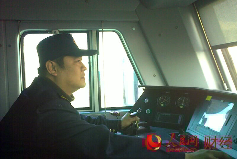 A man in uniform operates a train inside the driver's cabin, focused on the controls. Sunlight comes through the window. The People.cn Finance logo is in the bottom right corner.