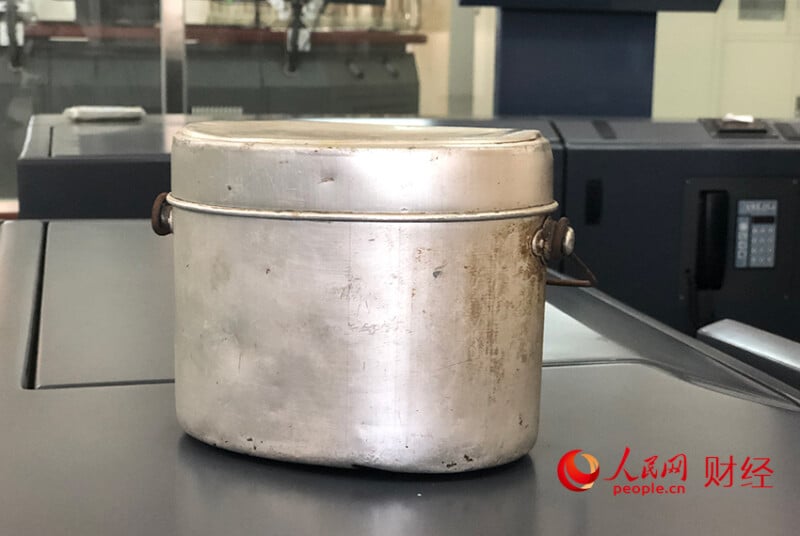 A worn, metal lunch tin with a lid and side latches sits on a dark surface. The container shows signs of use and age. The logo and text "people.cn" appear in the lower right corner.