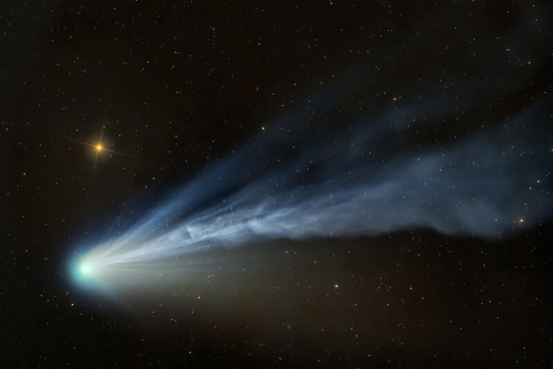 A bright comet with a glowing nucleus and a long, blue-tinged tail streaks across a star-filled night sky. The tail fans out and appears wispy against the dark backdrop of space.